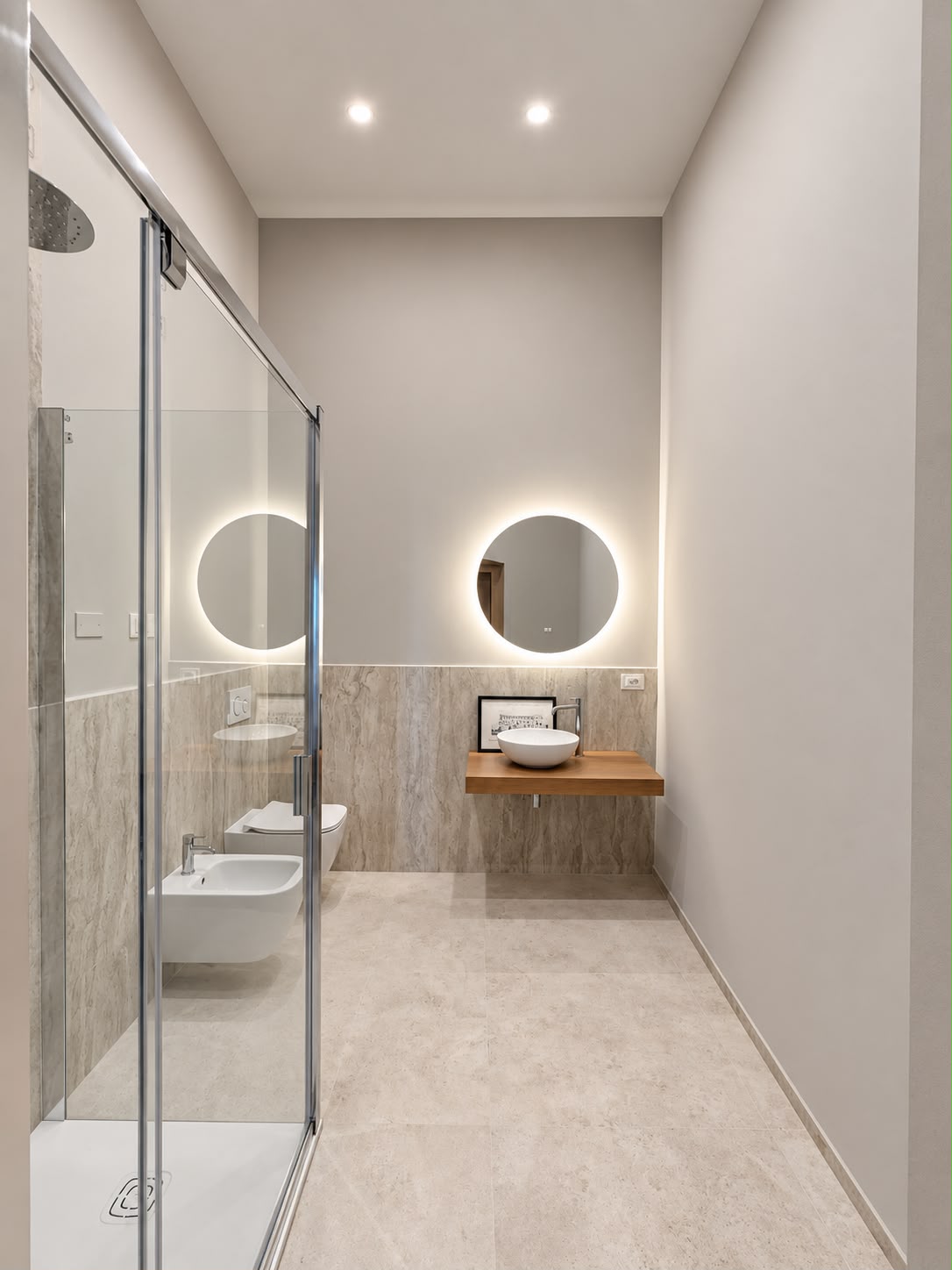 Bagno ristrutturato con box doccia in cristallo, doppio lavabo e rivestimenti in marmo chiaro.