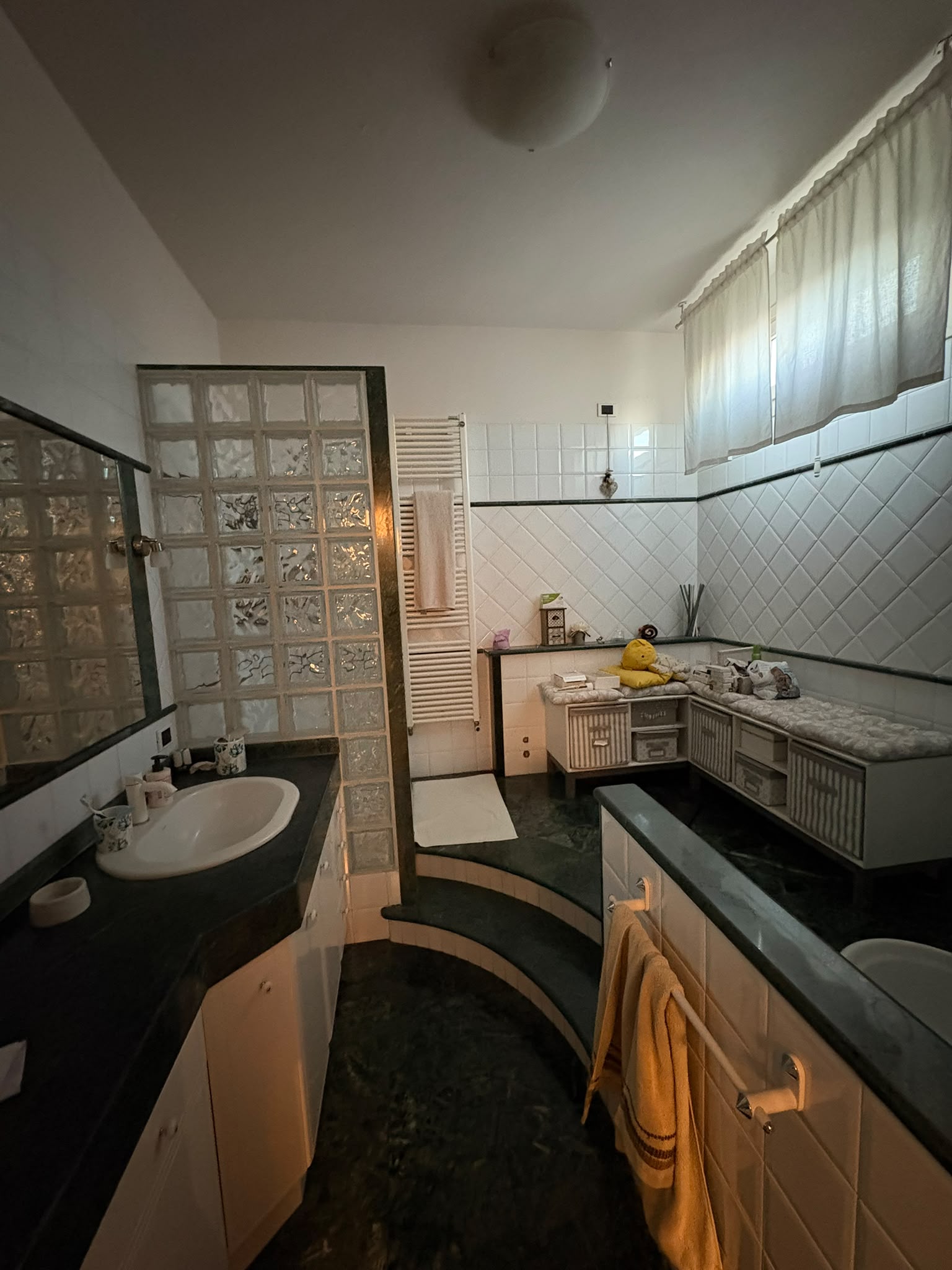 Bagno rivestito in marmo verde con doccia, due lavabi, termoarredo e parete in vetrocemento.