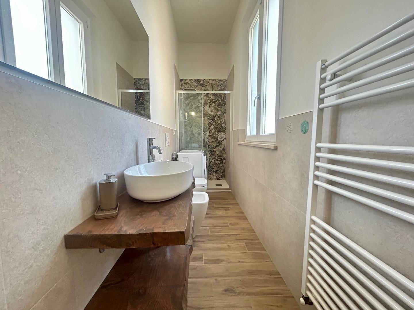 Bagno con doppia finestra, lavabo d'appoggio su mensola in legno, box doccia e scaldasalviette.