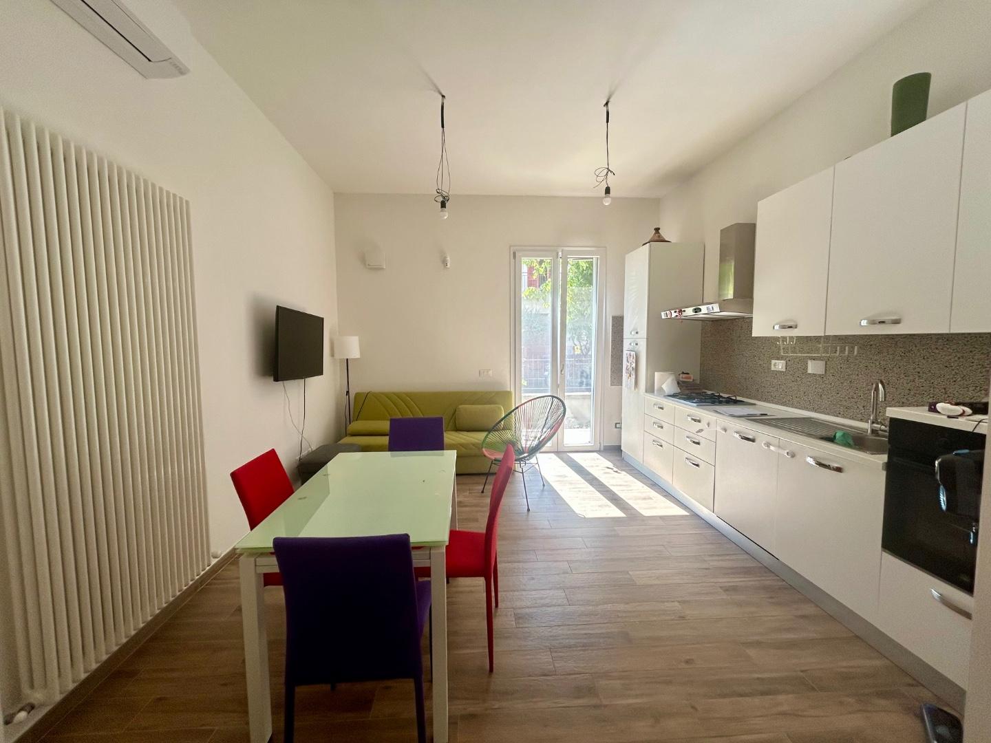 Vista d'insieme della zona living con tavolo da pranzo, cucina a vista e climatizzatore, pavimento in gres effetto legno.